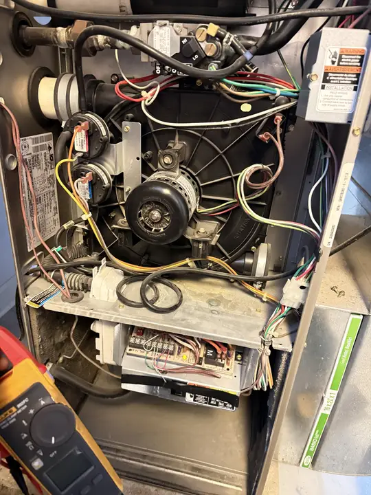 Inside a gas furnace showing the blower, inducer motor, pressure switches and control board, with a clamp meter in the foreground