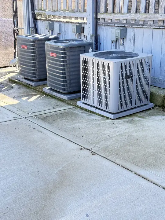 Three air conditioner condensers on concrete pads beside a wooden fence, two Goodman and one Ameristar